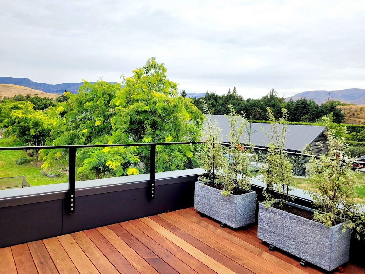A spacious deck is outlined by black railing, featuring planters that hold greenery. The view includes lush trees and rolling hills, creating a calming outdoor setting. The wooden deck boards provide a warm contrast to the greenery surrounding the area.