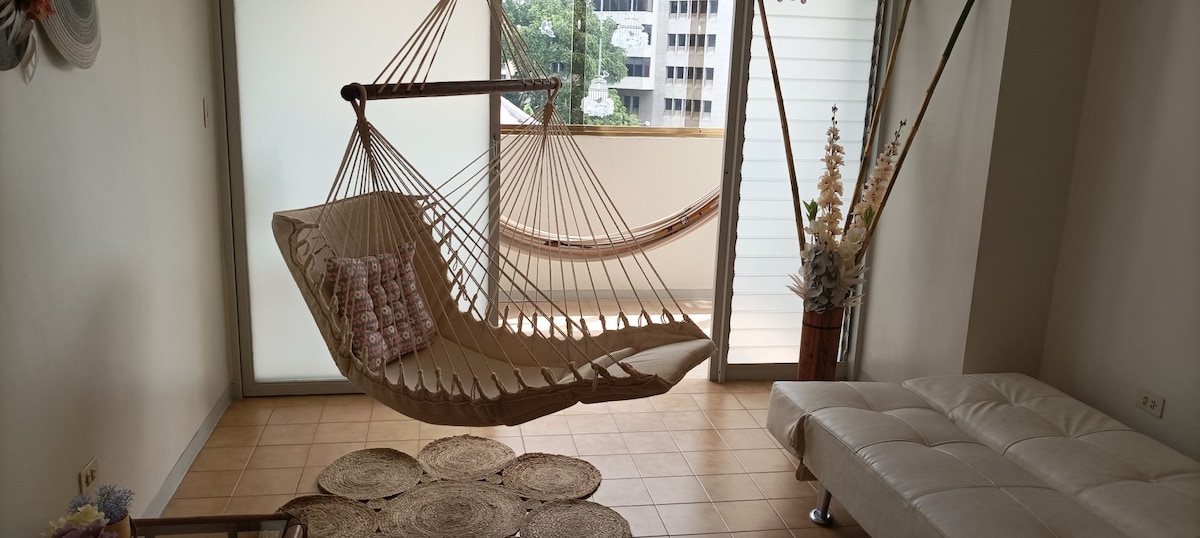 A comfortable sitting area features a hanging hammock and a stylish white sofa. Natural light filters through large glass doors that open to a balcony, enhancing the space. A round woven rug adds texture to the tiled floor, with a few decorative plants positioned nearby.