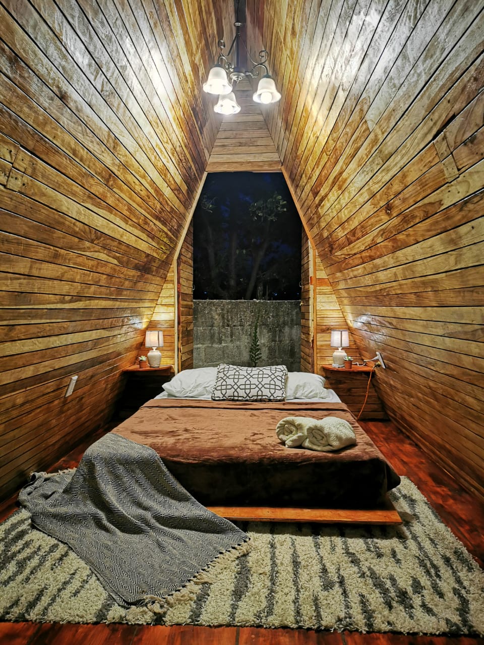 The interior features an A-frame structure with wooden paneling, complemented by a warm, inviting color palette. A spacious bed is centered below a window, allowing natural light to fill the space. Nightstands with lamps are positioned on either side, creating a cozy atmosphere.