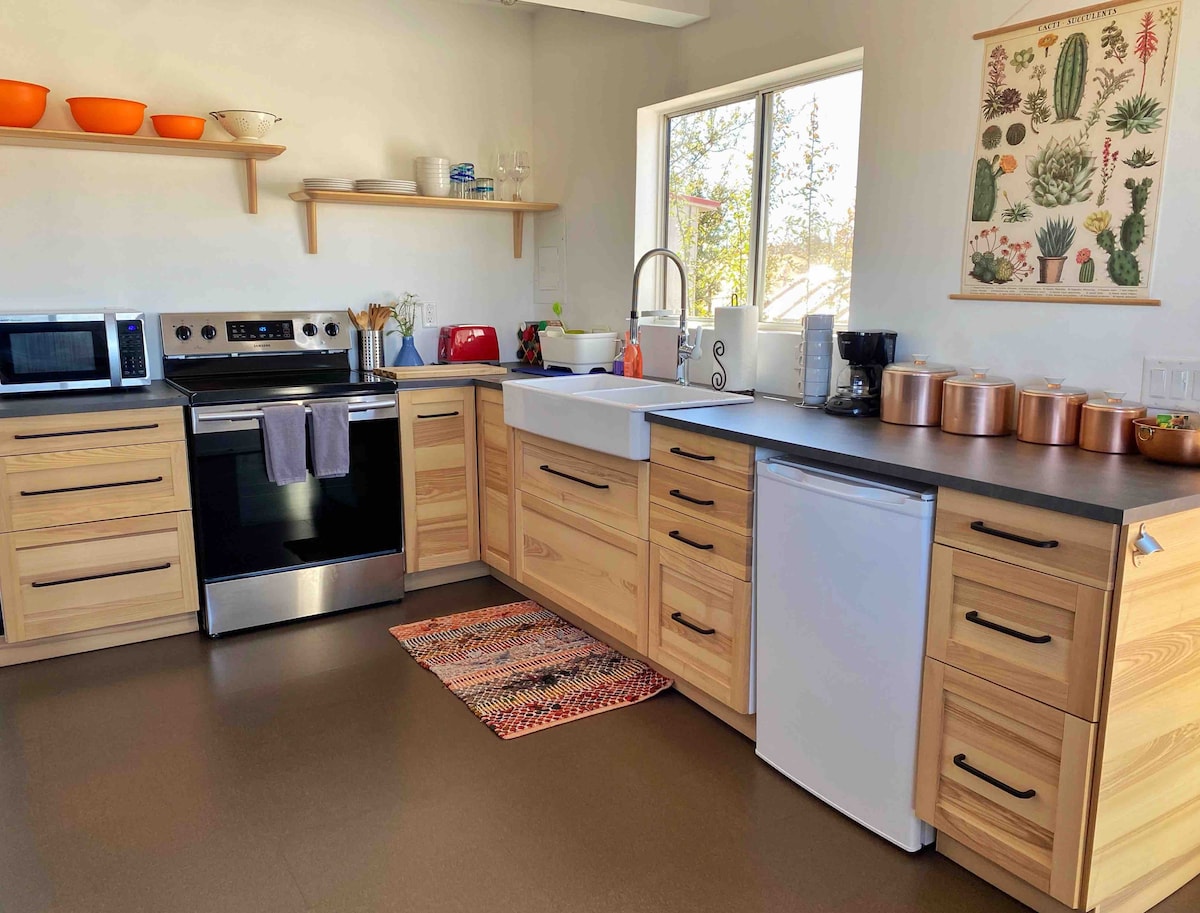 The kitchen features modern wooden cabinetry and a large window that brings natural light into the space. Appliances include a refrigerator, oven, microwave, and coffee maker. Counter space is complemented by utensils and dishware, with a decorative rug adding warmth to the floor.