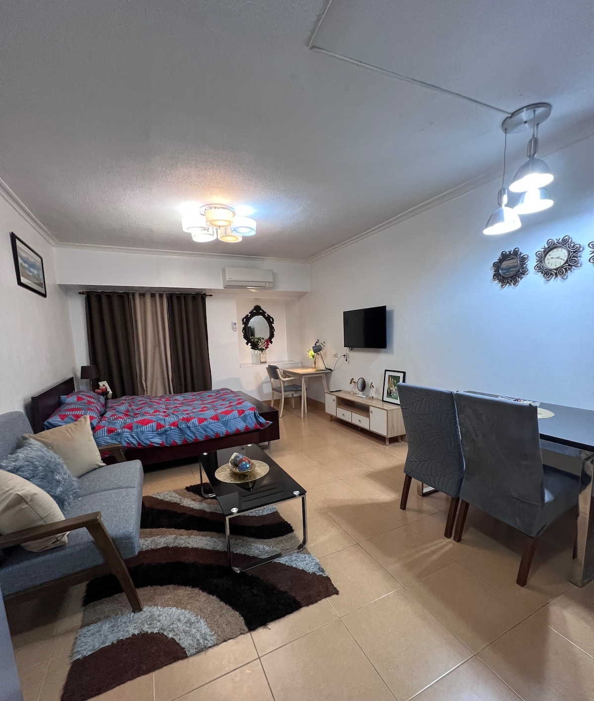 A fully furnished studio features a double bed adorned with complete bedding. A cozy couch is positioned nearby, while a dining table provides space for meals. A flat-screen TV is mounted on the wall, and natural light is filtered through floor-length curtains.
