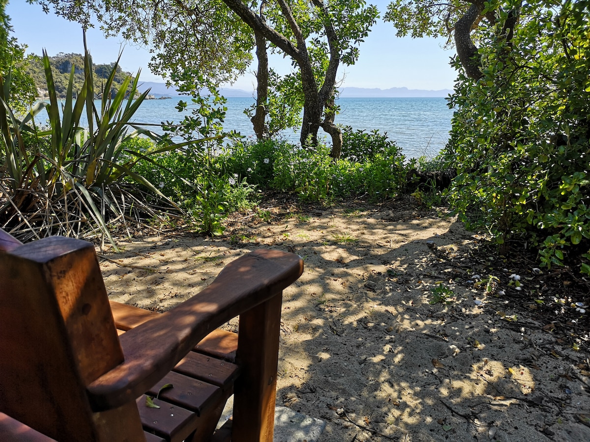 A serene view of the beach is framed by lush greenery and trees. A wooden chair is positioned on a sandy area, inviting relaxation while overlooking the water. The landscape features a blend of sunlight and shade from the surrounding foliage.