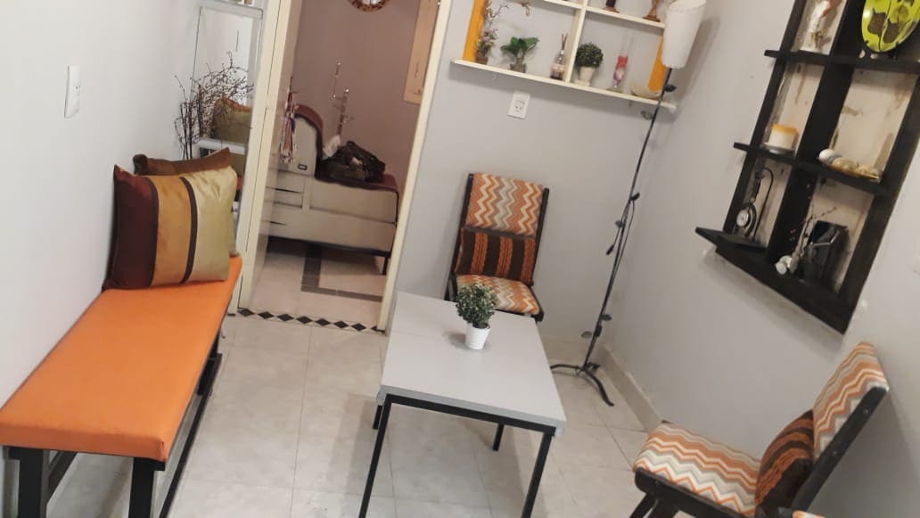A cozy seating area features two patterned chairs and a bench, all in warm orange tones. A small light-colored table sits in the center, adorned with a potted plant. A shelving unit displaying decorative items is mounted on the wall, contributing to the inviting ambiance.