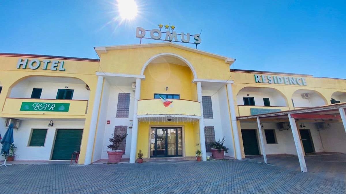 A yellow building displays a welcoming entrance, featuring a large archway and dual signage indicating 'Hotel' and 'Residence.' Potted plants line the entry, and bright sunlight casts a warm glow, creating an inviting atmosphere.