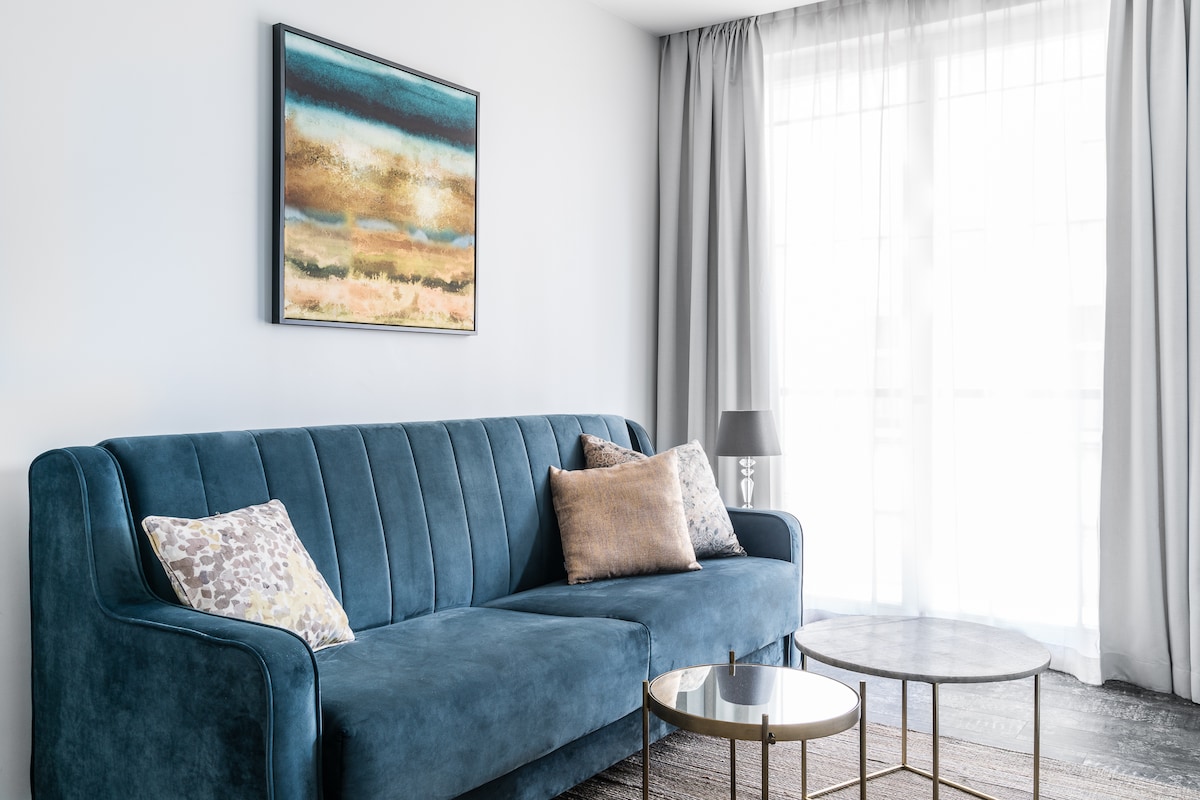 A stylish living area features a plush blue sofa adorned with decorative cushions. A sleek coffee table sits in front, with a piece of abstract artwork displayed on the wall above. Natural light filters through sheer curtains, creating a serene and bright ambiance.