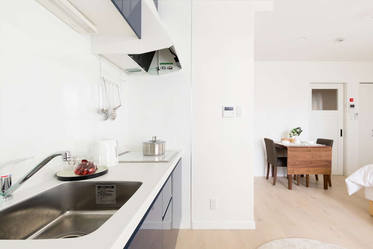 A well-equipped kitchen area features a stainless steel sink and sleek countertops. Cooking utensils are neatly arranged above, while a saucepan is visible on the stove. In the background, a dining table with chairs is softly illuminated, creating a welcoming dining space.