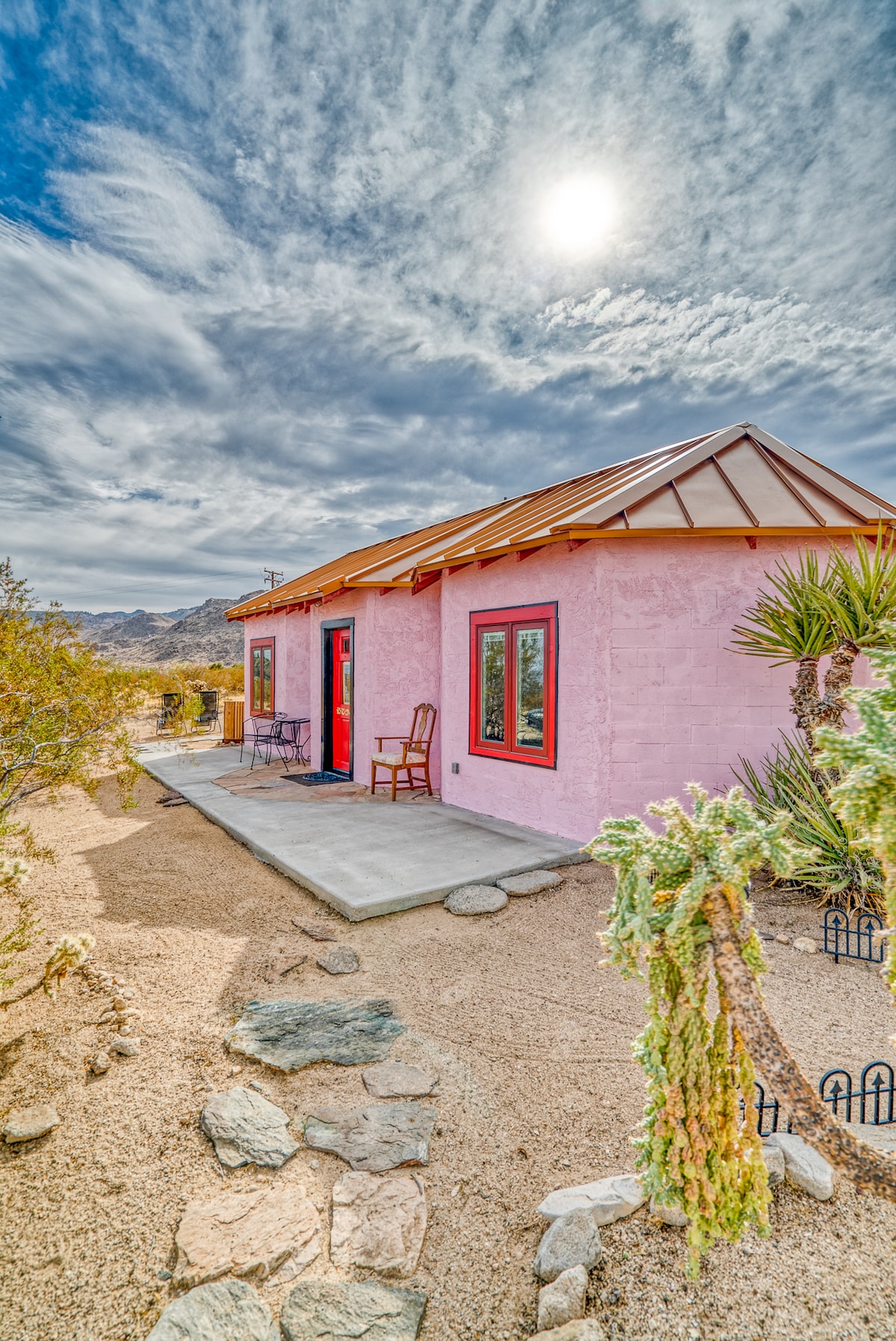The exterior of a pink cabin is featured, surrounded by desert landscaping. A small seating area with a chair and a table is visible on the patio, along with various plants. Rocky ground textures complement the vibrant structure under a partly cloudy sky with the sun shining brightly.