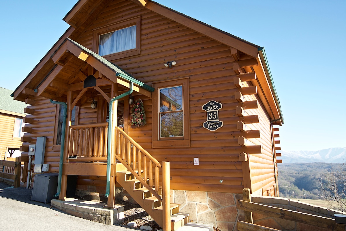 The rustic exterior of the cabin is showcased with warm wooden siding and a welcoming front porch. A large wreath is displayed on the door, and the cabin number is prominently featured. Scenic mountain views can be seen in the background under a clear blue sky.