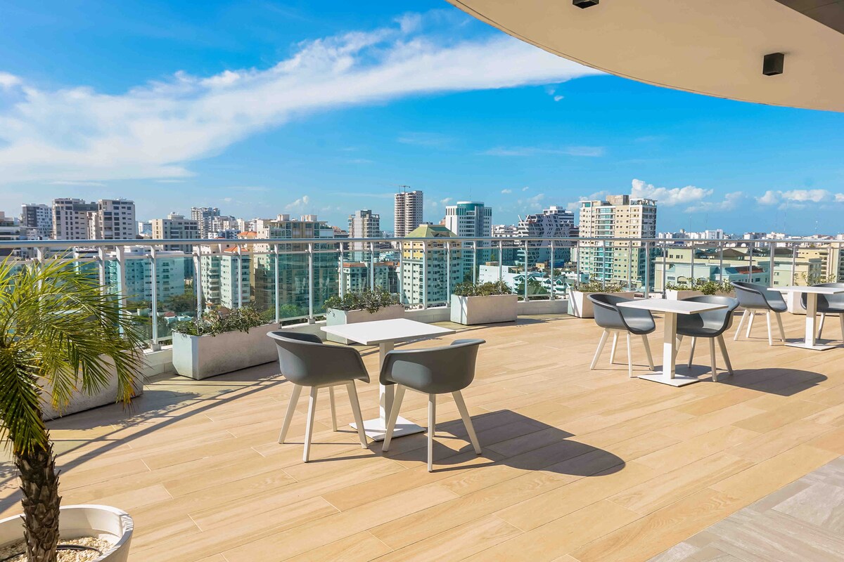 A spacious outdoor terrace provides sweeping views of the city skyline. Multiple tables and chairs are arranged for seating, with greenery and planters enhancing the ambiance. The clear blue sky complements the modern architecture that surrounds the area.