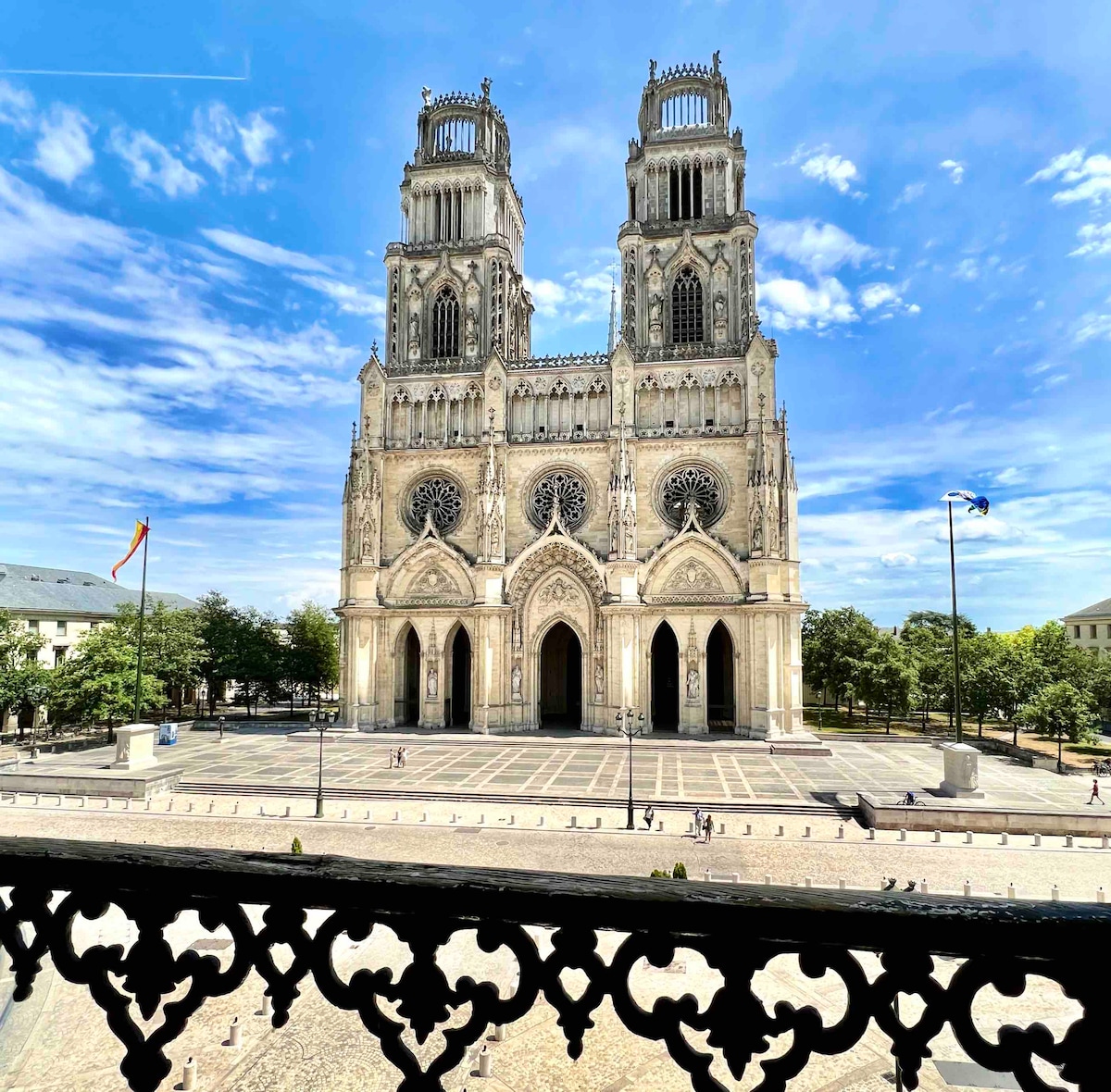 The image captures the stunning façade of the Cathedral, featuring intricate gothic architecture and towering spires. In the foreground, the view includes a decorative balcony railing. The vibrant sky and sunlit square below enhance the scene.