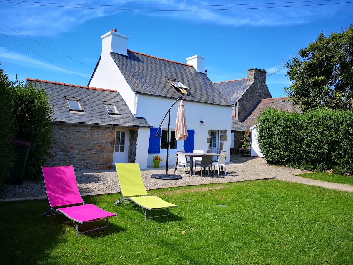 A renovated house is showcased, featuring blue shutters and a slate roof. The outdoor space includes a manicured lawn with two lounge chairs, one in pink and the other in green. A patio table and umbrella are visible, surrounded by neatly trimmed hedges.