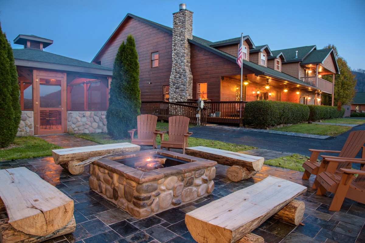 An outdoor seating area is centered around a stone fire pit, featuring wooden benches and Adirondack chairs. The rustic building is visible in the background, with a stone chimney and a neatly landscaped yard, contributing to a cozy, communal atmosphere.