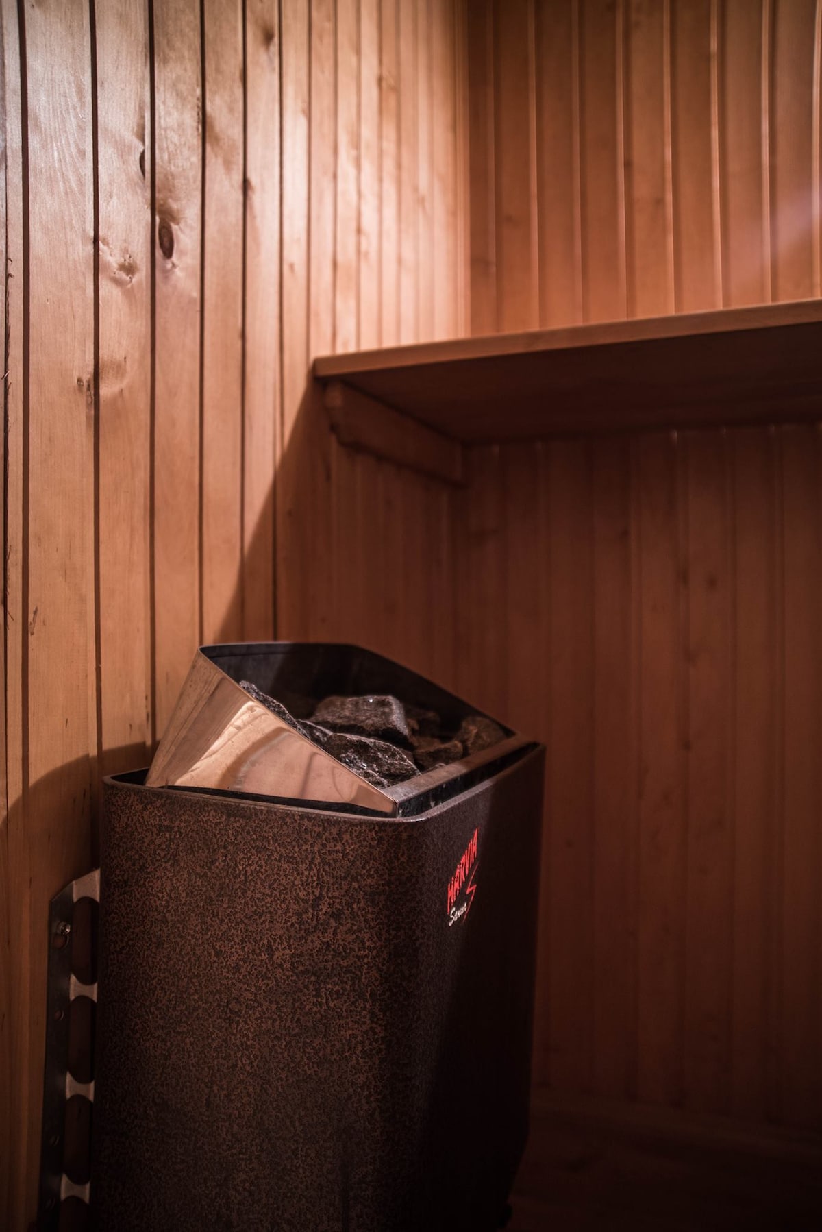 A sauna heater with a metal reservoir for stones is positioned against a wooden wall clad in light wood paneling. A small shelf is visible above the heater, providing additional storage or display space. The warm tones of the wood enhance the relaxing environment.
