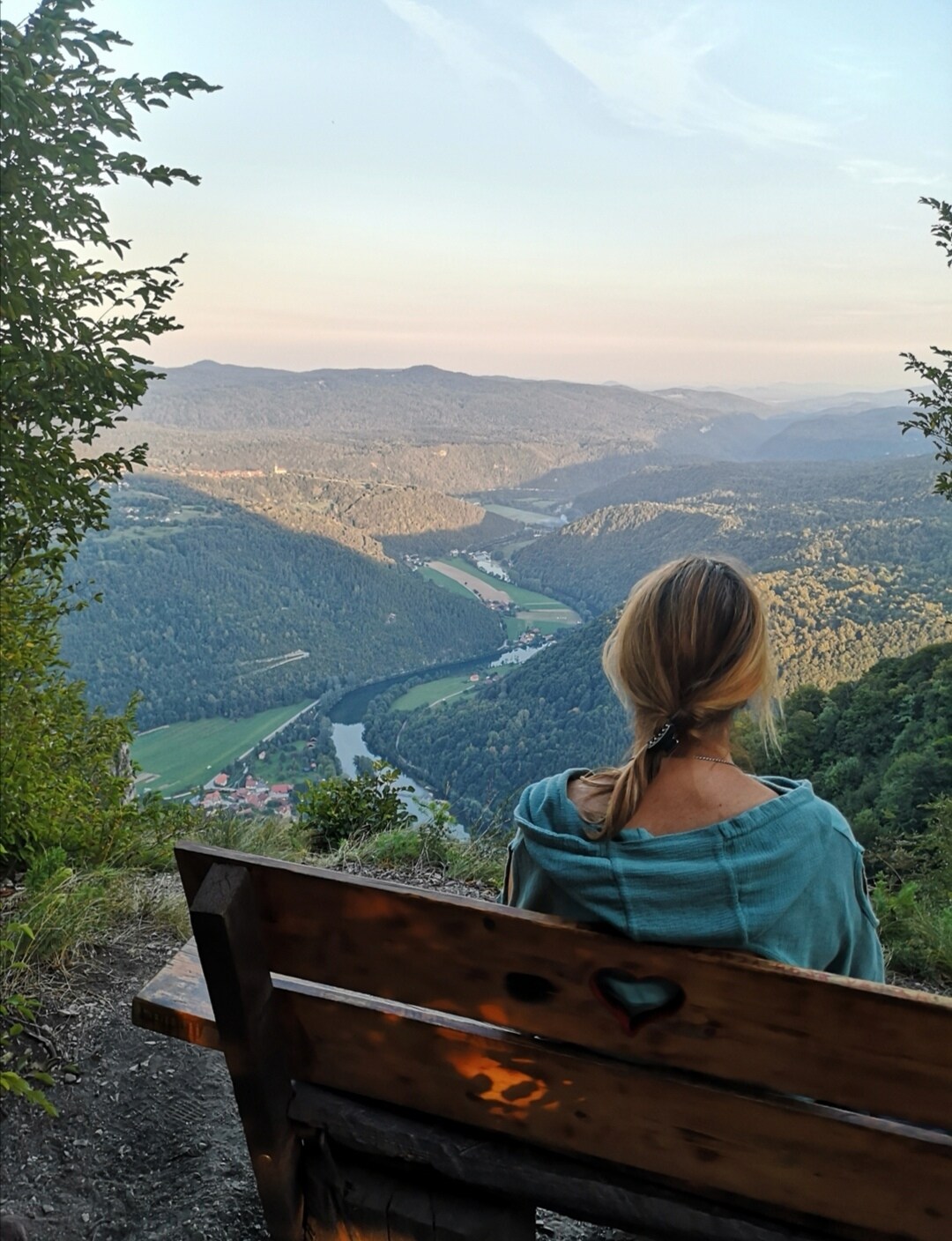 A person seated on a wooden bench overlooks a sprawling valley. Rolling hills and a winding river are visible below, surrounded by lush greenery. The soft hues of the sunset cast a warm glow over the landscape, creating a sense of tranquility.