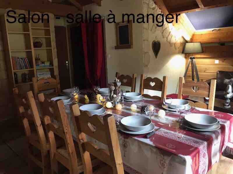 A large dining area is furnished with a long wooden table surrounded by chairs. The table is set with plates and decorative items, creating a welcoming environment. Wooden shelves filled with books are visible in the background, contributing to the cozy ambiance.