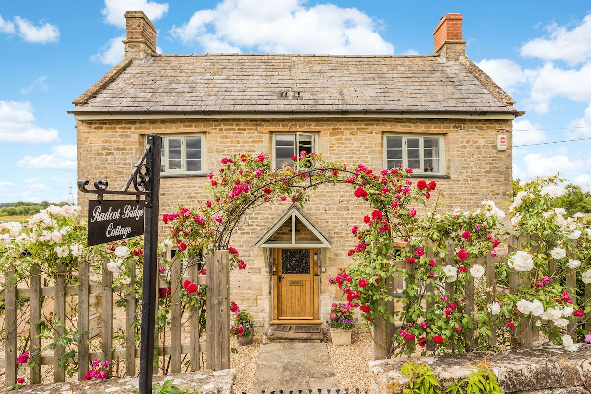 The cottage exterior is framed by vibrant flowers in full bloom, enhancing the charm of the stone façade. A welcoming wooden door serves as the centerpiece, while the name 'Radcot Bridge Cottage' is displayed on an elegant sign. Clear blue skies provide a serene backdrop.