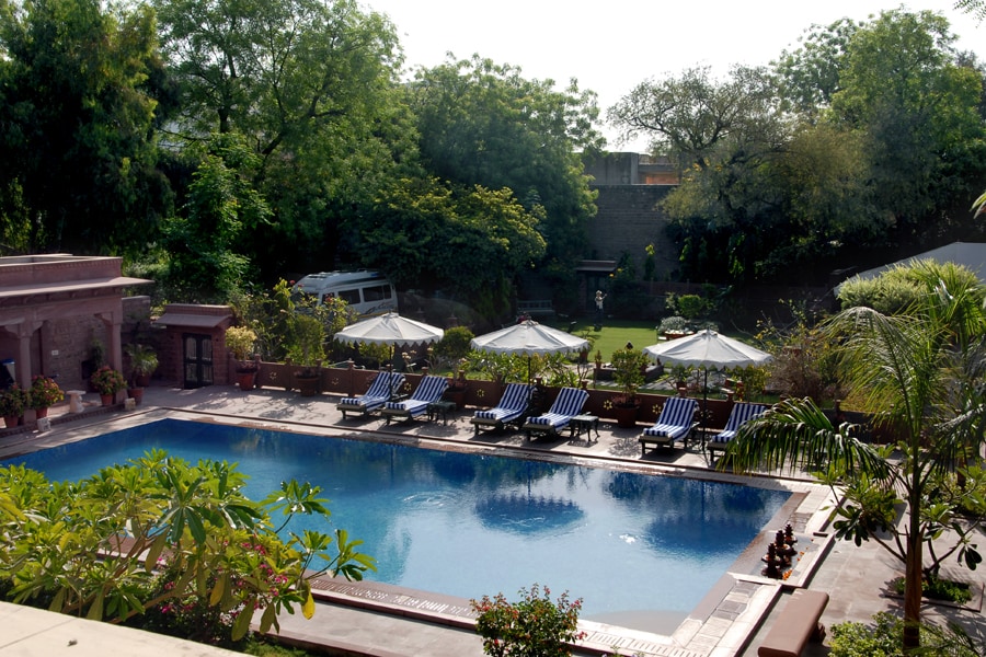 A serene pool area features lounge chairs with striped cushions, surrounded by lush greenery. Umbrellas provide shade over some seating, while the clear water of the pool invites relaxation. The spacious garden in the background offers a peaceful setting for guests to enjoy.