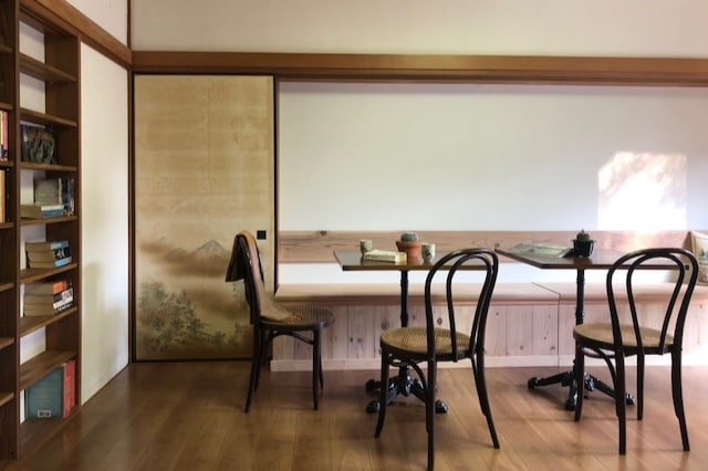 An inviting interior space showcases a minimalist design featuring two wooden tables with stylish chairs. A wall-mounted bookshelf displays neatly arranged books, and a sliding door adorned with a mountain motif complements the serene atmosphere.
