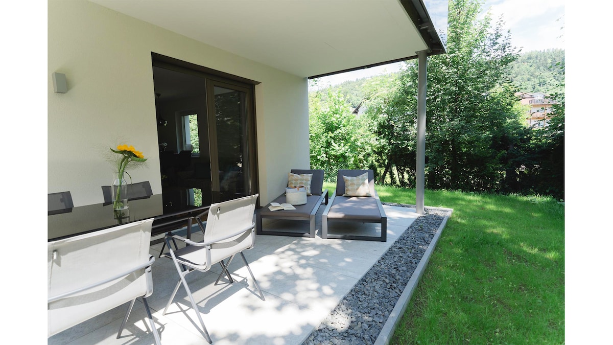 A covered terrace features a dining table with chairs and two lounge chairs positioned for relaxation. Lush greenery surrounds the space, providing a natural view. The concrete patio is complemented by pebbled borders, enhancing the outdoor setting.