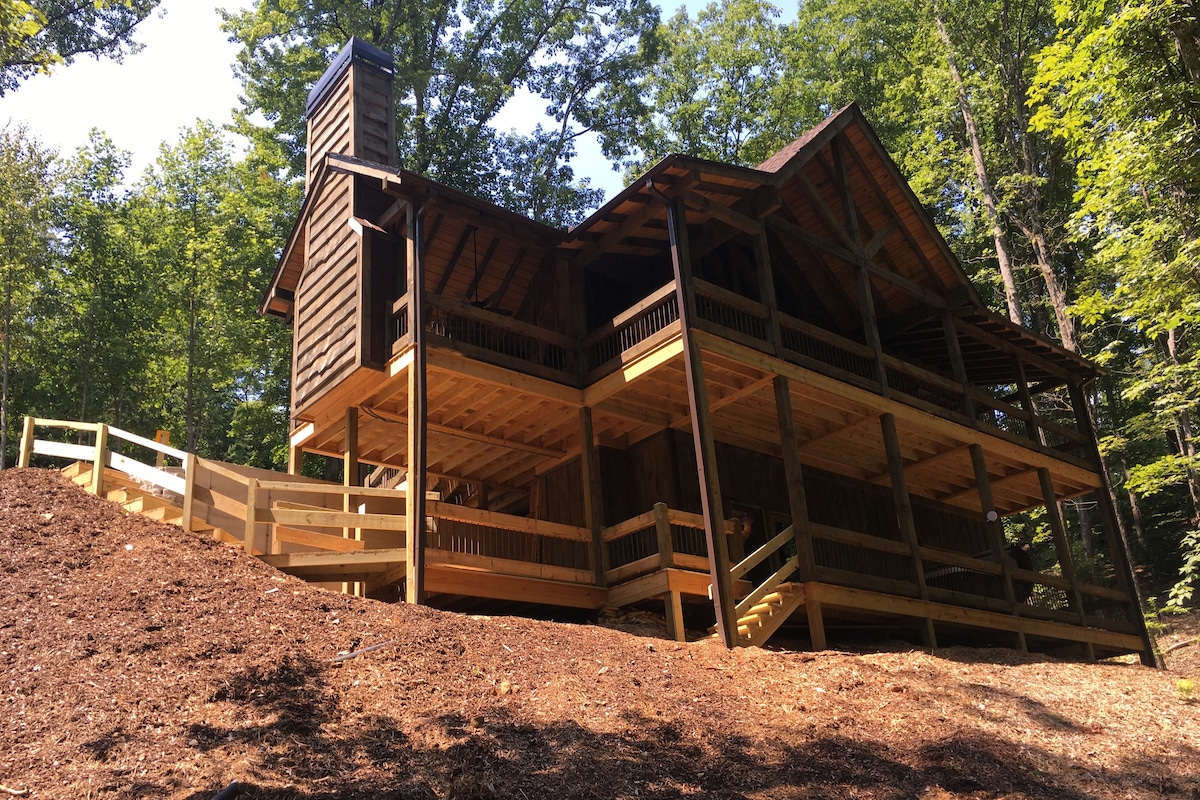 A newly constructed cabin showcases a rustic exterior with a combination of wooden siding and a stone chimney. Surrounded by trees, the structure features a covered deck on two levels and a well-maintained path leading to the entrance.