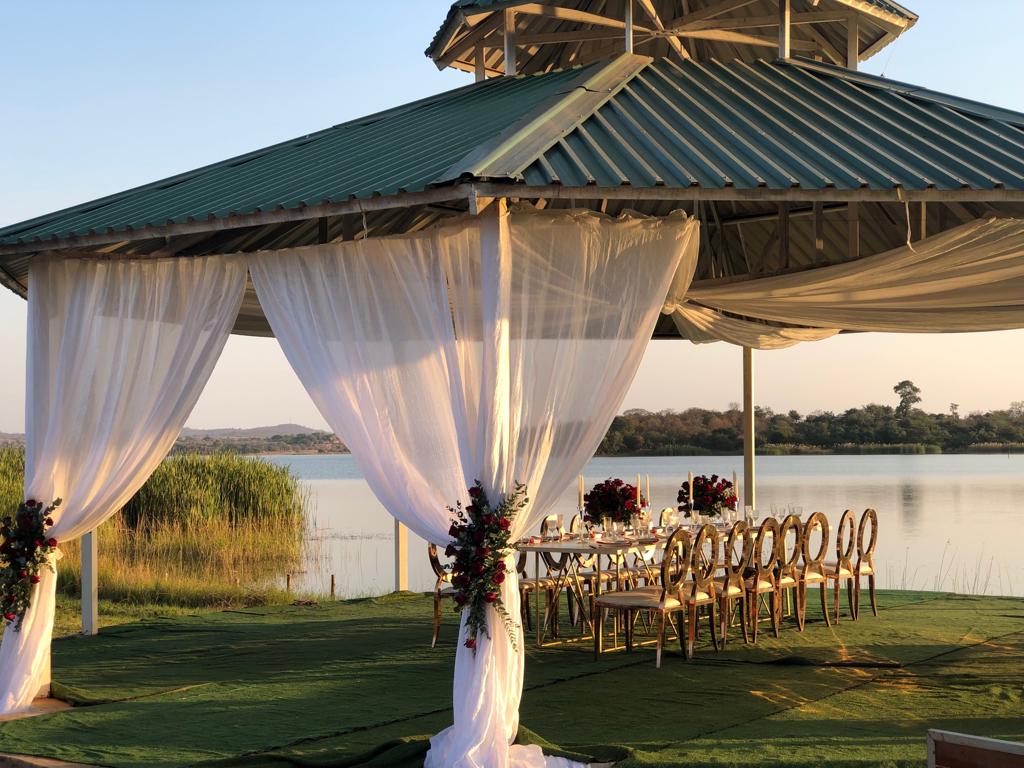 An outdoor dining area is shaded by a large gazebo featuring a green roof. Elegant chairs surround a long table, adorned with floral decorations. Soft, sheer curtains frame the entrance, while scenic views of the waterfront and lush greenery provide a tranquil backdrop.