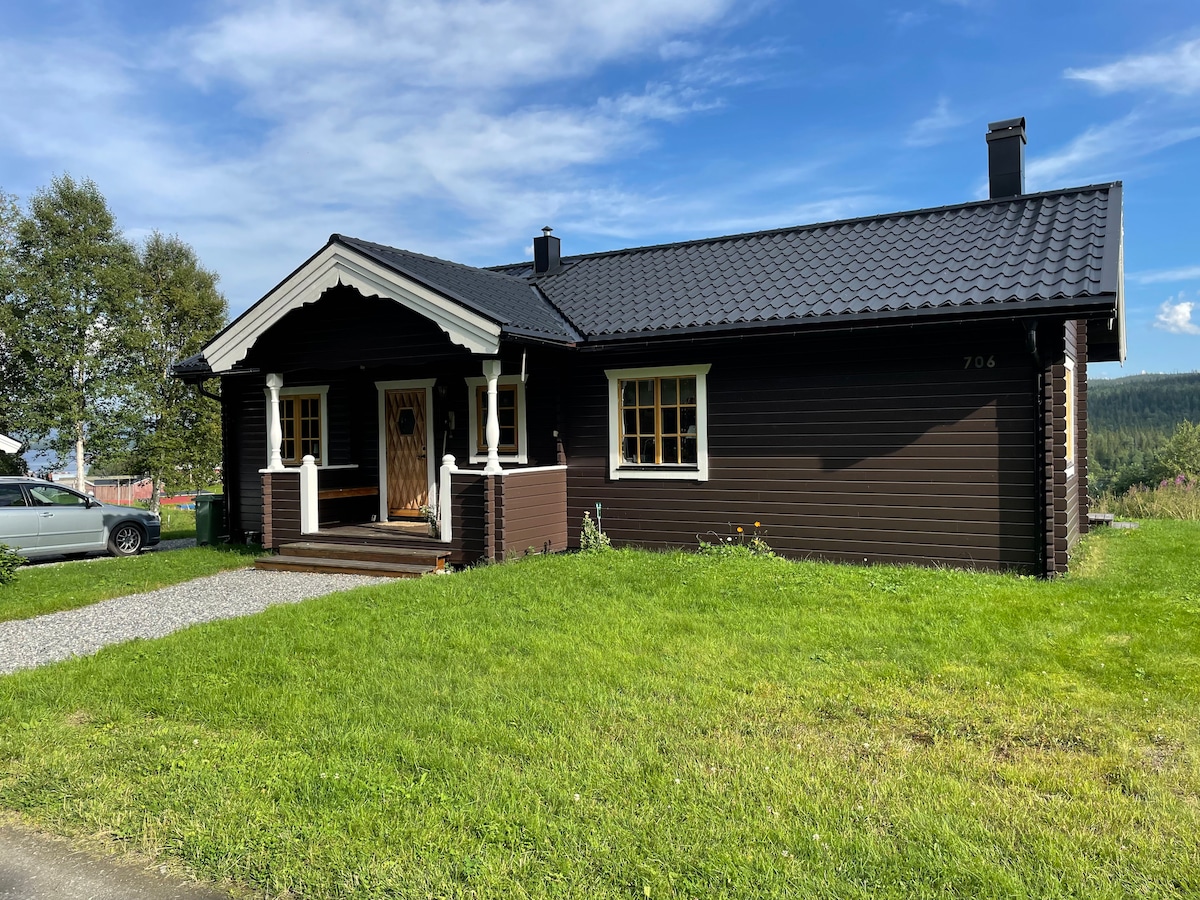 A charming wooden cabin features a dark exterior with white trim accents. A welcoming entrance is framed by traditional architecture, while a neat pathway leads through a well-maintained lawn. The surrounding landscape displays a serene setting with greenery and distant hills under a clear blue sky.