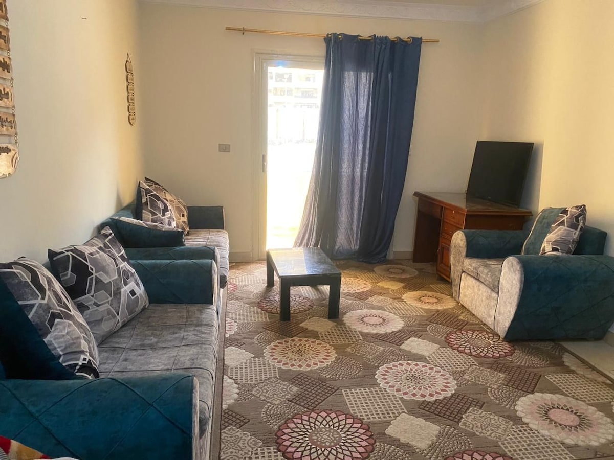 A well-furnished living area features a combination of textured sofas and armchairs arranged around a central coffee table. A large window draped with blue curtains allows natural light to fill the space. A TV is placed on a wooden cabinet against one wall.