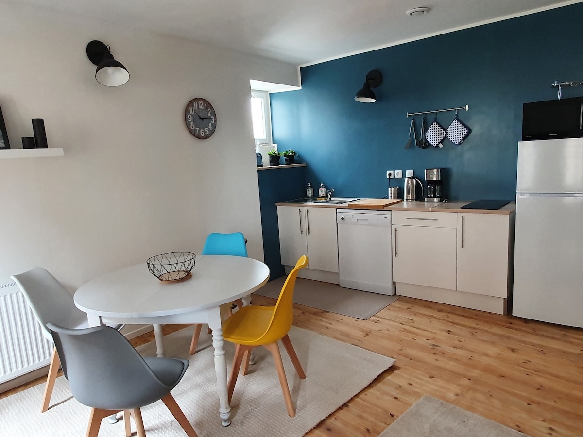 A modern kitchen and dining area feature a round dining table surrounded by four chairs in varying colors. The blue wall accentuates the space, while a clock and kitchen utensils are displayed. A dishwasher and refrigerator add functionality to the well-lit area.
