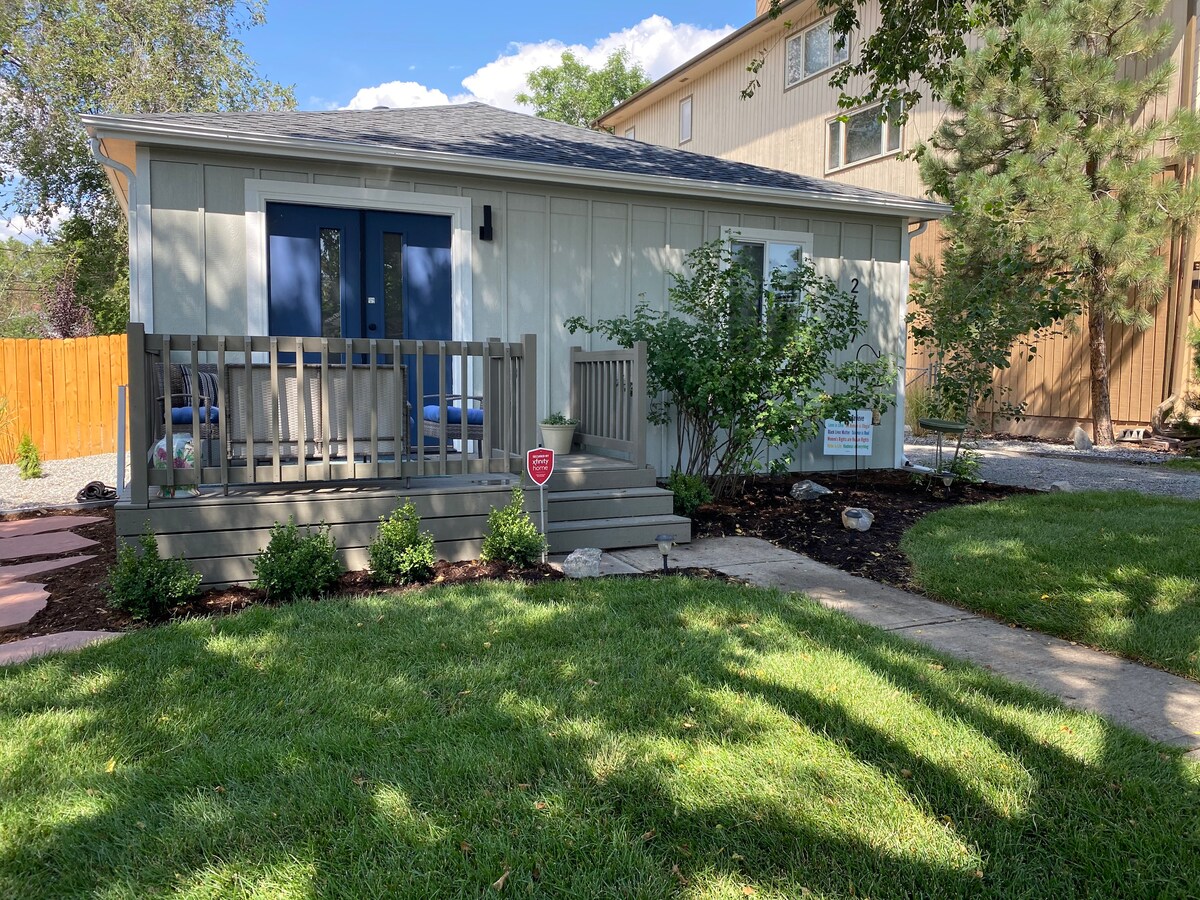 A light-colored home is framed by vibrant green grass and new landscaping. The front porch features wooden railings and steps leading to double blue doors. A welcoming sign is displayed near the entry, surrounded by small plants and shrubbery.