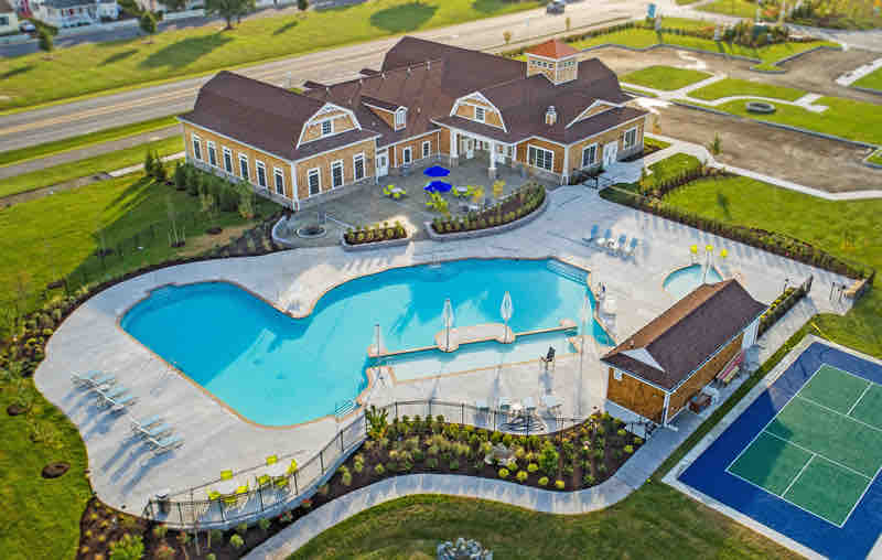 An aerial view showcases a grand community center with a large, irregularly shaped pool surrounded by lounge chairs and umbrellas. A quaint pool house is visible nearby, along with a tennis court outlined on the property. Lush landscaping encircles the recreational areas.