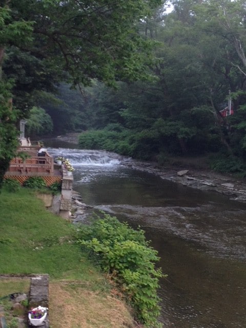 A serene view of 16 Mile Creek flows gently alongside the cottage, framed by lush greenery. A wooden deck is positioned nearby, providing a peaceful space to enjoy the natural surroundings. The sound of water adds to the tranquil atmosphere.