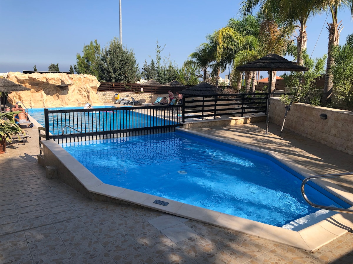 A communal swimming pool area is visible, featuring a sparkling blue pool bordered by stone tiles. Palm trees provide shade, along with thatched umbrellas placed around the poolside, while a separate shallow section is designed for relaxation and leisure.