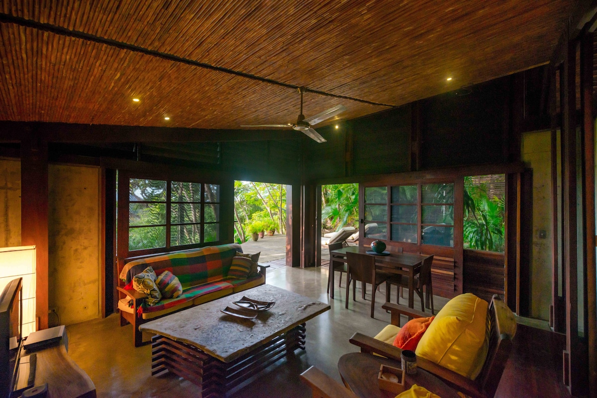An open living area features a colorful sofa and a natural rock coffee table. Large sliding walls provide views of the lush outdoor surroundings. Ceiling fans circulate air beneath the wooden ceiling, enhancing comfort in the spacious environment.