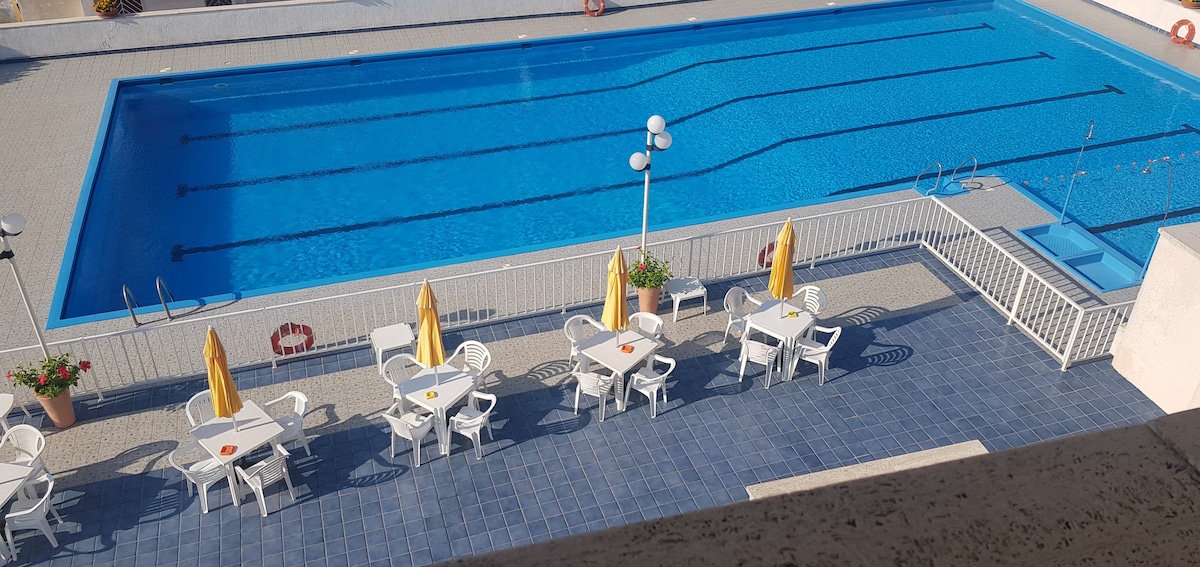 A large swimming pool is visible, surrounded by tiled flooring and adorned with sun umbrellas. Several white chairs and small tables are set up nearby, creating a space for relaxation. The area is well-lit, and the pool is marked with lanes for swimming.