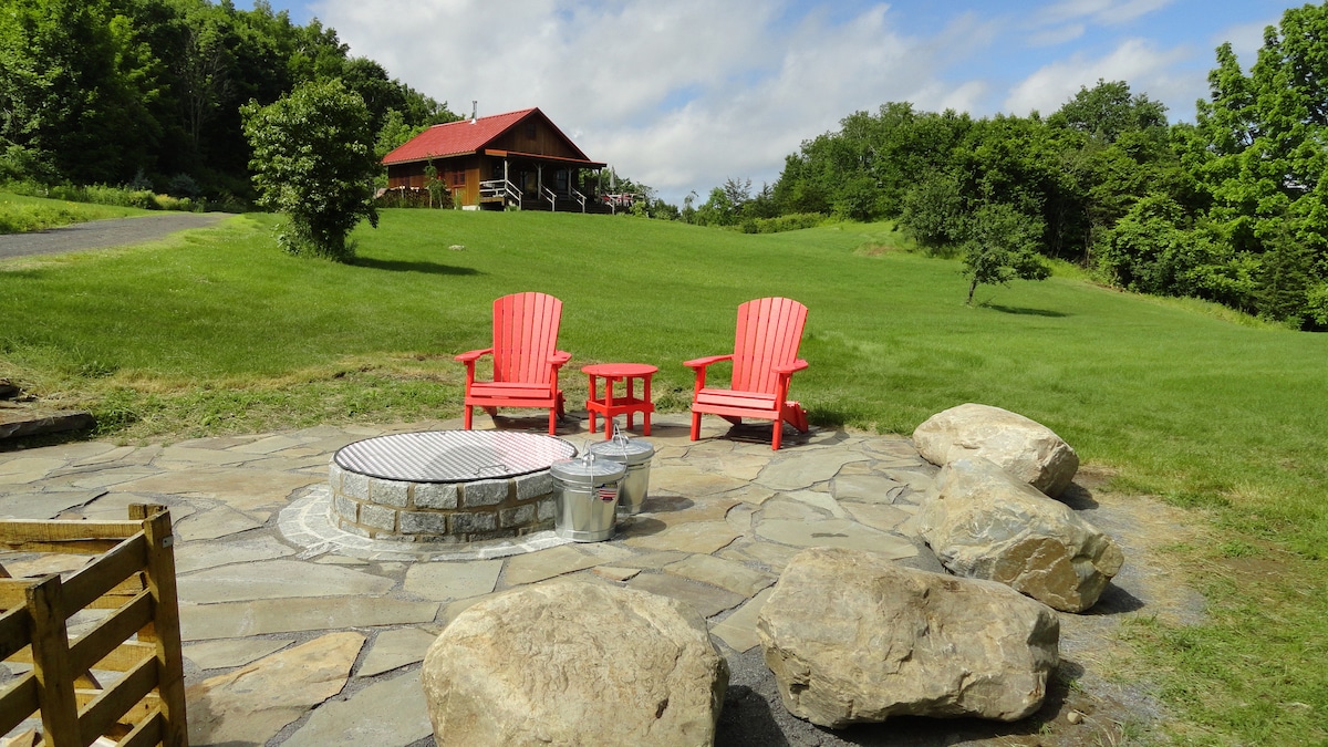 An outdoor fire pit area is surrounded by large, flat stones, with two red Adirondack chairs and a small table positioned nearby. Lush green grass leads to a distant wooden cabin under a partly cloudy sky, enhancing the serene landscape.
