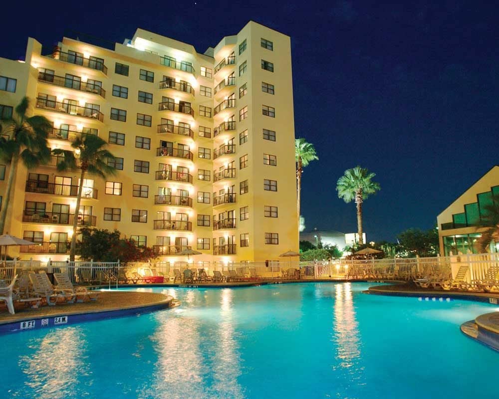 A well-lit, multi-story building is seen at night, surrounded by palm trees and reflecting lights in an inviting pool. Lounge chairs are neatly arranged, and the pool area is illuminated in soft blue tones, creating a calm atmosphere.