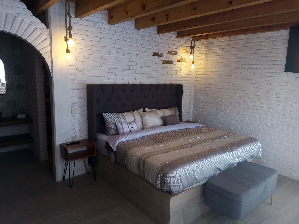 A spacious room features a king-size bed adorned with decorative pillows and a subtle blanket. Warm lighting is provided by hanging fixtures above the bed, while a bedside table complements the wooden beams of the ceiling. A cozy bench is positioned at the foot of the bed.