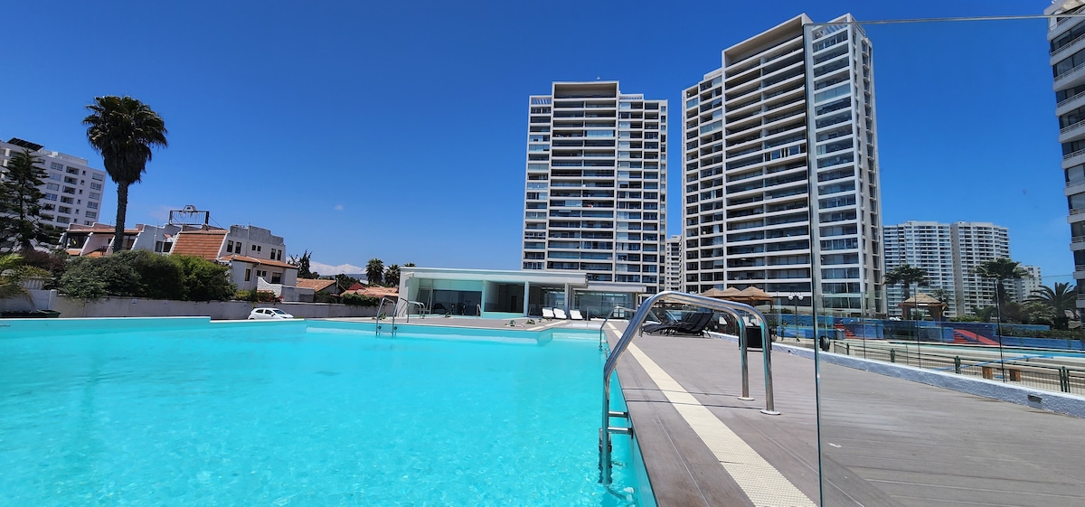 A modern swimming pool is featured, reflecting bright blue water under a clear sky. Surrounding modern buildings are visible in the background, and a deck area lined with loungers is situated beside the pool, inviting relaxation and leisure.