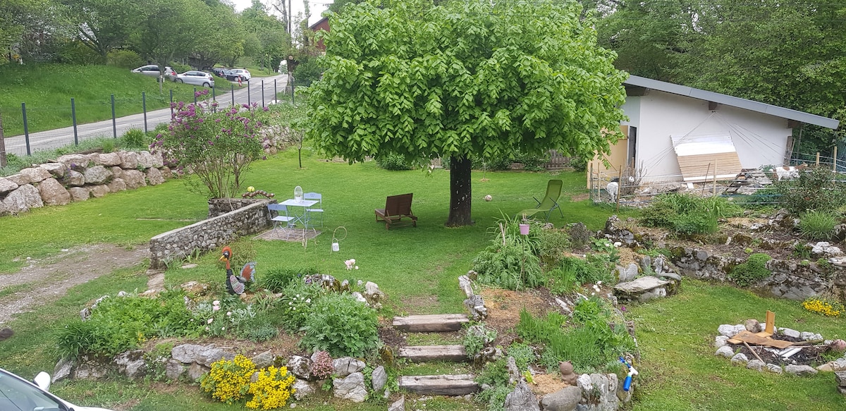 A spacious garden features a large green lawn with a variety of trees and plants. Stone steps lead through flower beds to a shaded area with a table and chairs. Surrounding features include a gravel path, decorative stones, and a glimpse of the nearby parking area.