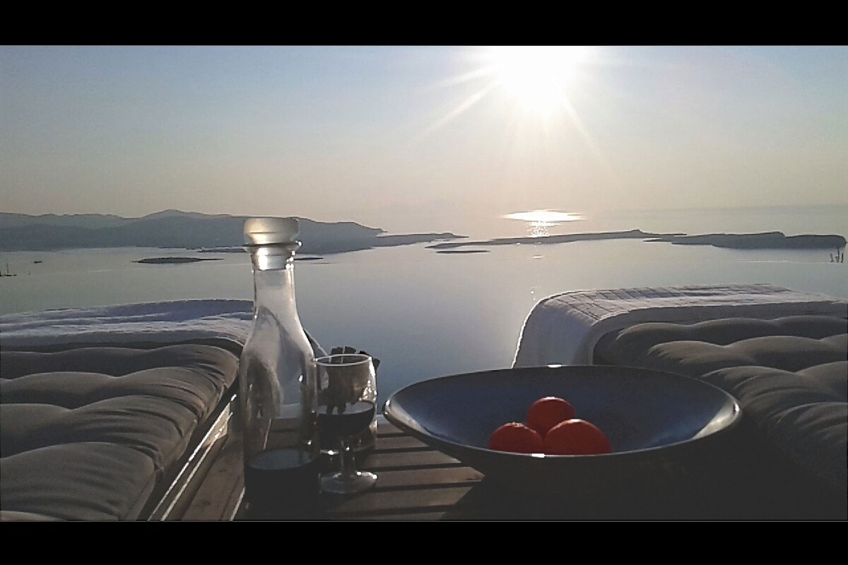 A tranquil scene captures the sun rising over a calm sea, with distant islands visible on the horizon. A glass of red wine and a carafe rest on a wooden table, accompanied by a bowl of red fruits. The spacious lounge area features comfortable cushions.