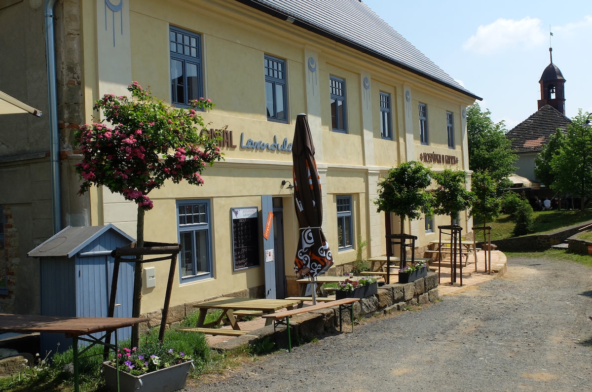 A charming building with a yellow exterior displays multiple large windows on the upper level. Outdoor seating is available on the gravel path, complemented by potted plants and vibrant flowering trees. A wooden structure with ornamental designs adds character to the inviting entrance.