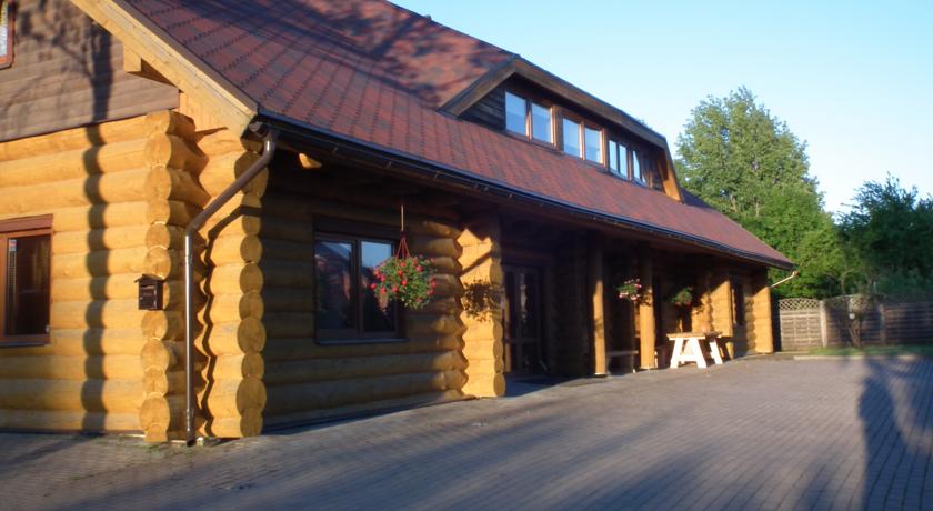 The exterior of the log house showcases natural wood construction with a sloped roof and a welcoming porch. Flower baskets adorn the entrance, while a broad pathway leads to the building, set against a backdrop of trees and clear blue skies.