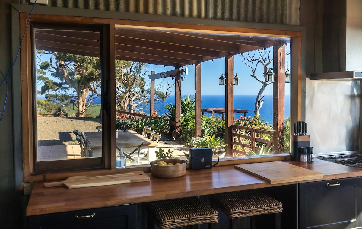 A country kitchen features wooden countertops and a bowl of yellow fruit. Large windows provide a scenic view of the outdoor dining area, surrounded by greenery and ocean vistas. Additional light fixtures hang above, enhancing the room's rustic charm.
