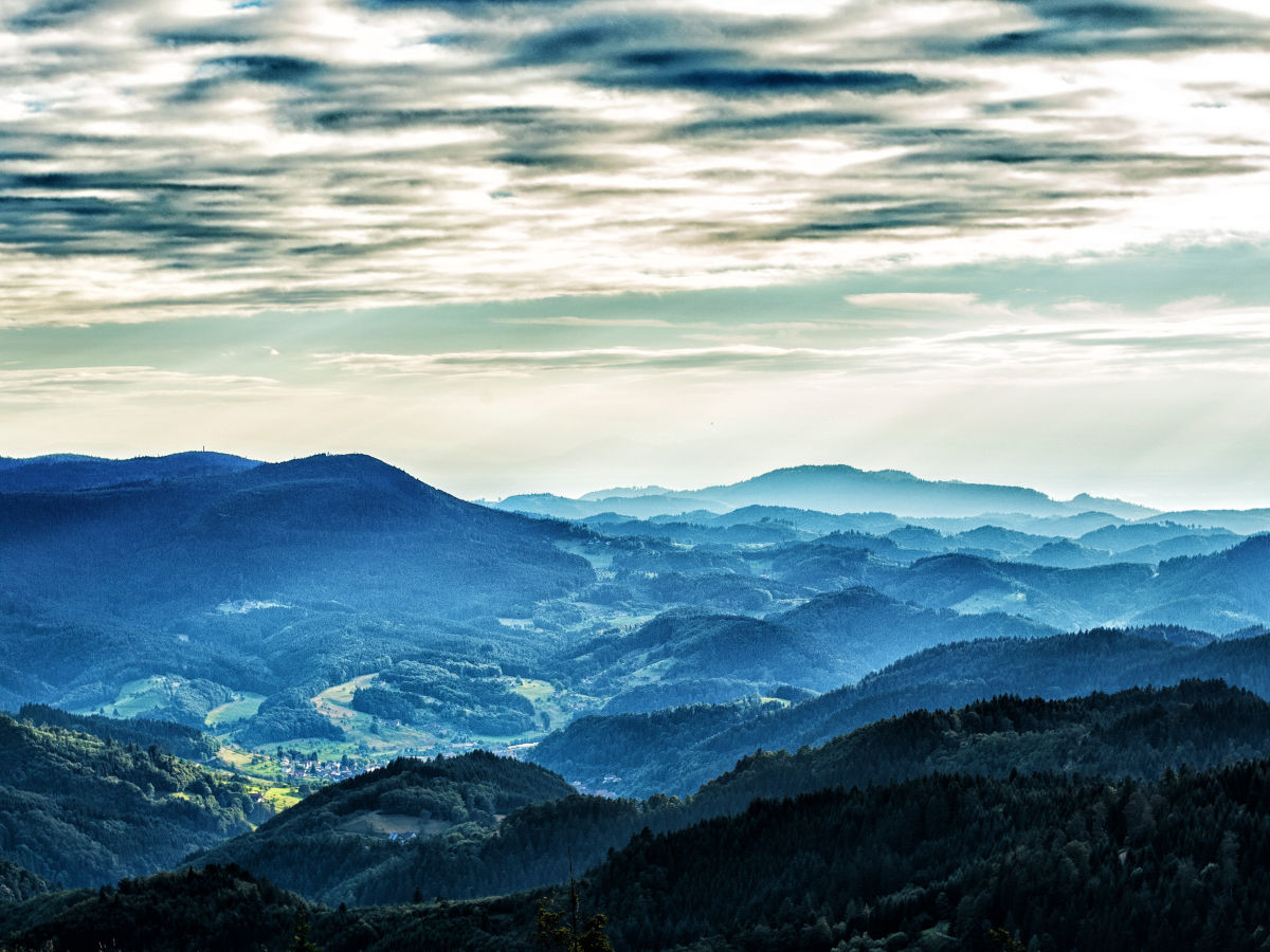 A scenic view of rolling hills and valleys under a partly cloudy sky is presented. Layers of blue and green hues create depth, as sunlight softly illuminates the mountains in the background, and a patchwork of fields can be seen in the foreground.
