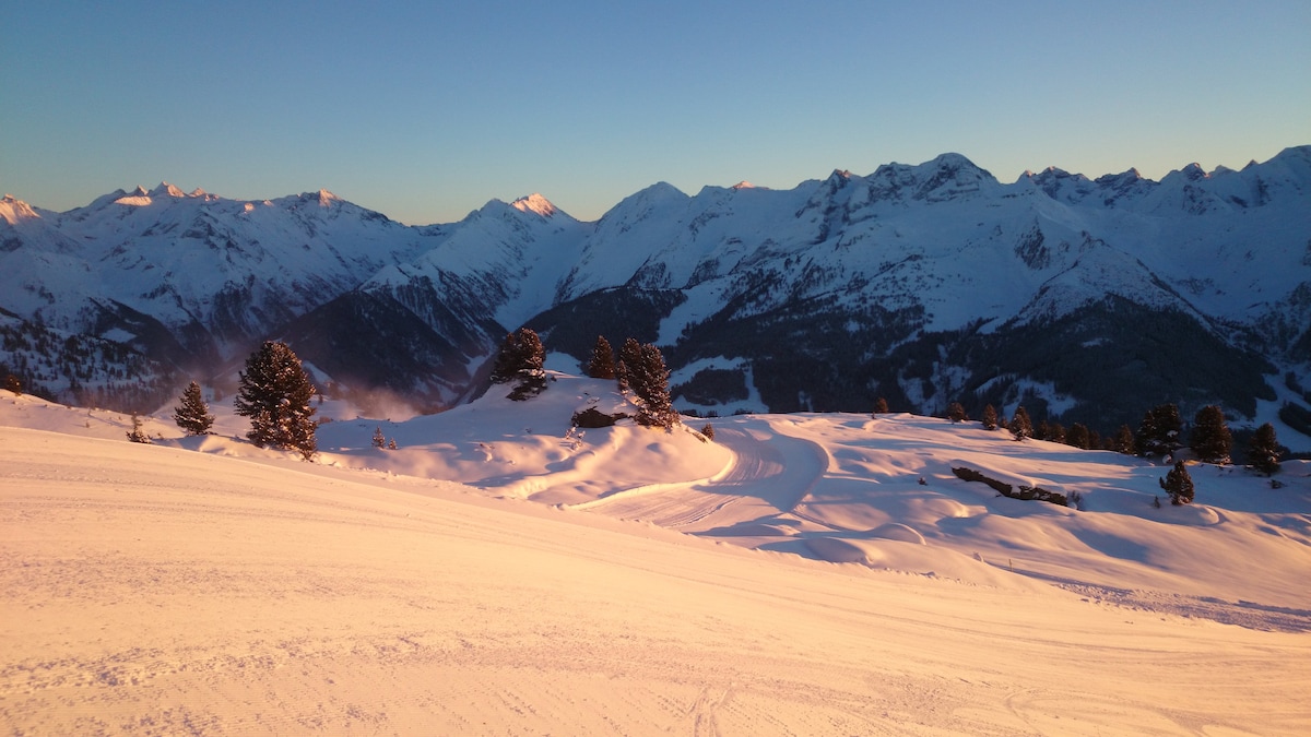 A panoramic view of a snow-covered mountain landscape is presented, with rolling hills and distant peaks bathed in the warm hues of sunrise. Tall trees punctuate the snow's surface, showcasing the natural beauty of the winter setting.