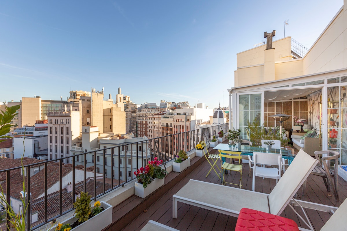 A spacious rooftop terrace features a variety of seating options, including lounge chairs and a glass-enclosed area. Planters with colorful flowers decorate the space, while a panoramic view of Madrid's skyline is visible in the background, showcasing a mix of historical and modern architecture.