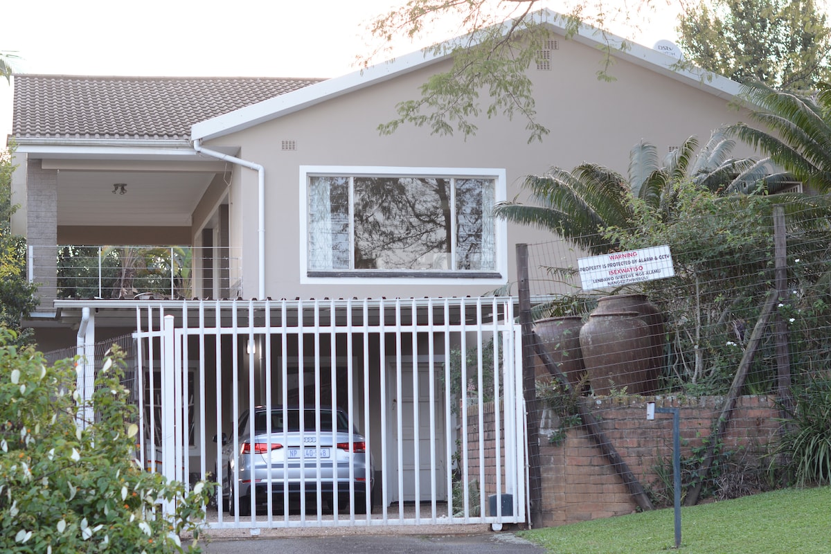A two-story home features a gated entrance with a white fence. A car is parked beneath the covered area on the ground level. Lush greenery is seen surrounding the property, with large pots and foliage adding to the inviting entryway.