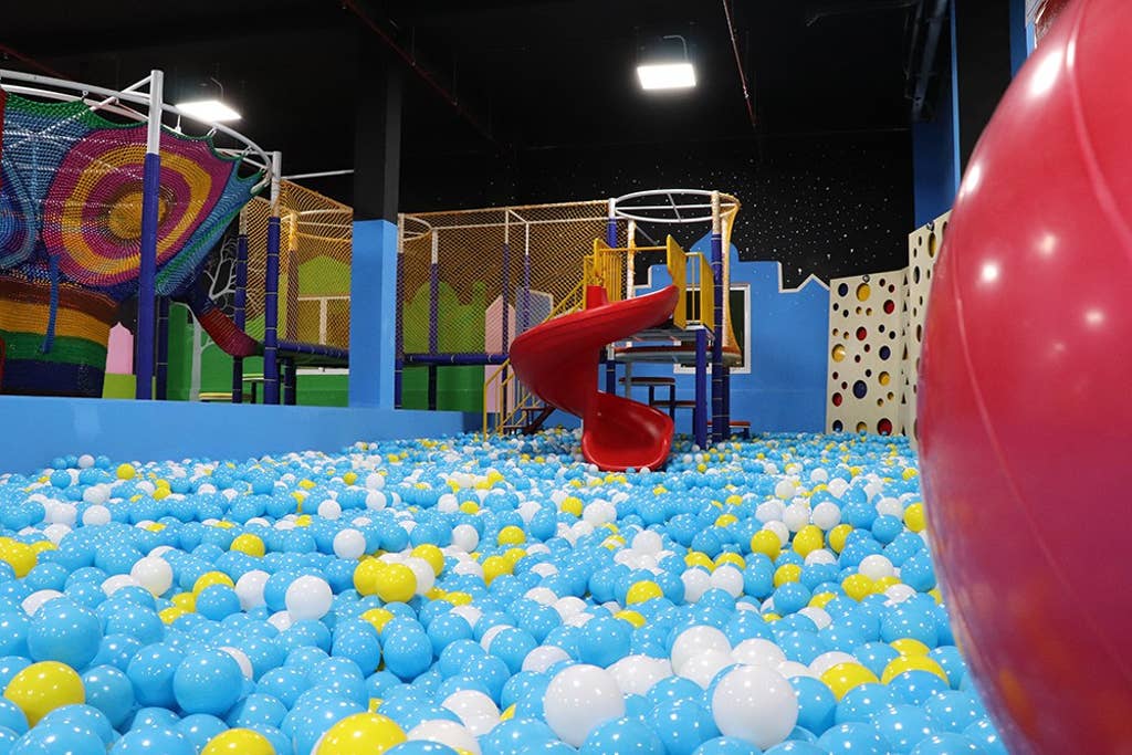 A vibrant play area is filled with colorful balls in shades of blue, yellow, and white. A red slide is positioned prominently, leading into the ball pit. Brightly colored structures and walls create a playful and engaging environment suitable for children.