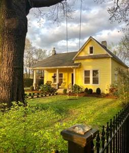 A charming yellow house with a covered front porch is set against a backdrop of green lawns and trees. The property is surrounded by well-maintained plants and is framed by an inviting iron fence. Two swings are visible on the porch and tree.