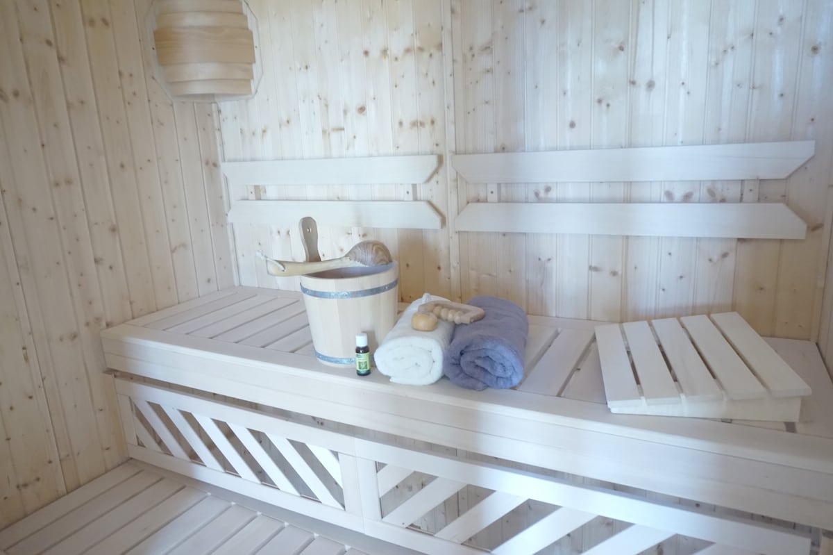 A wooden sauna interior features light-colored walls and benches. A wooden bucket and ladle are positioned next to soft towels. An aroma oil bottle is visible, providing a serene atmosphere suitable for relaxation.